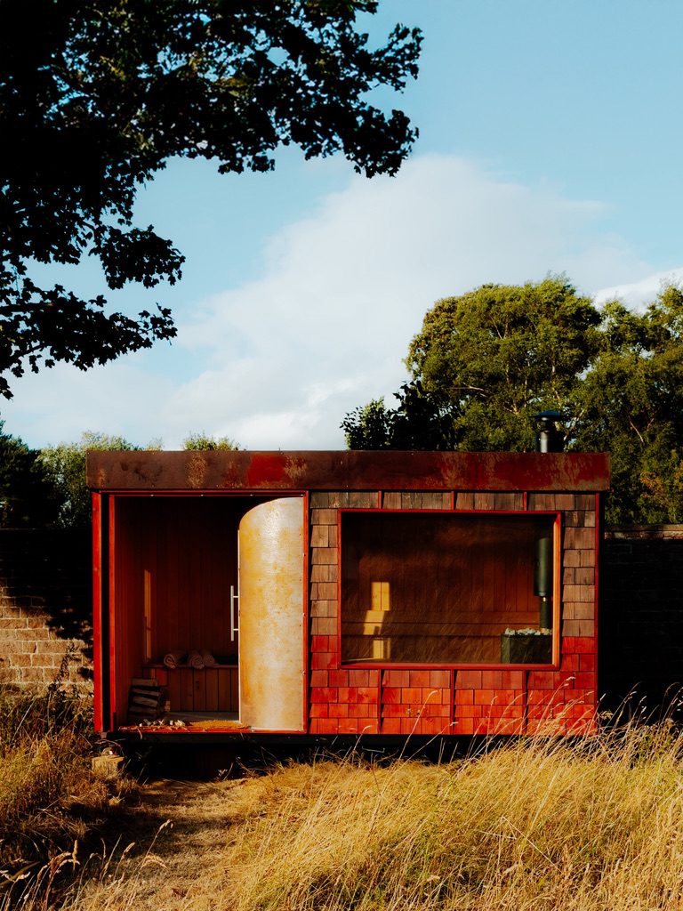 A Rigg mobile sauna in a wild garden — cedar shingle cladding, weathering-steel roof and a curved door open to the cedar-lined interior