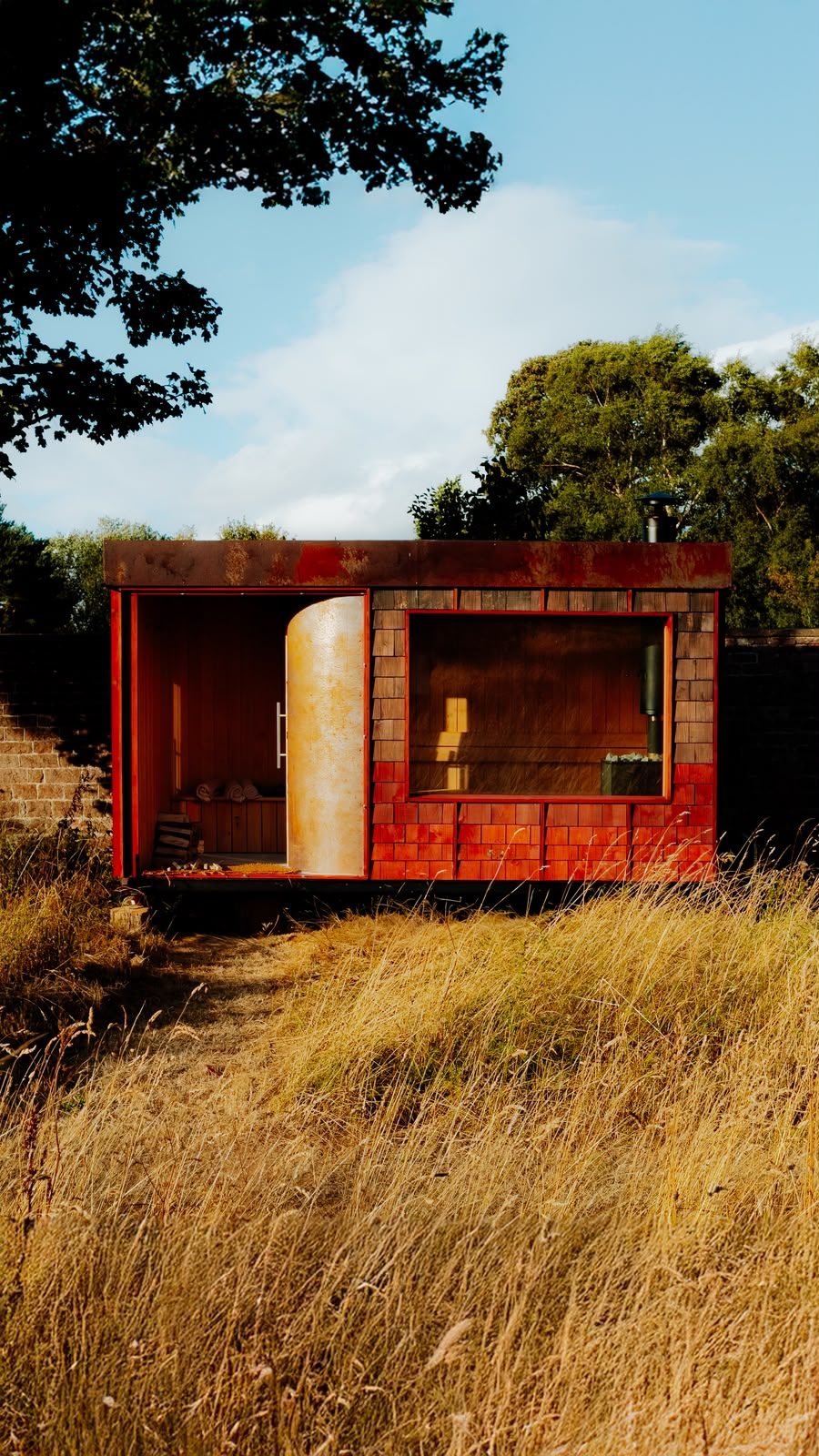 Rigg mobile sauna sited in a wild garden — western red cedar shingle cladding and a weathering-steel roof, its curved door open to the cedar-lined interior and wood-fired stove