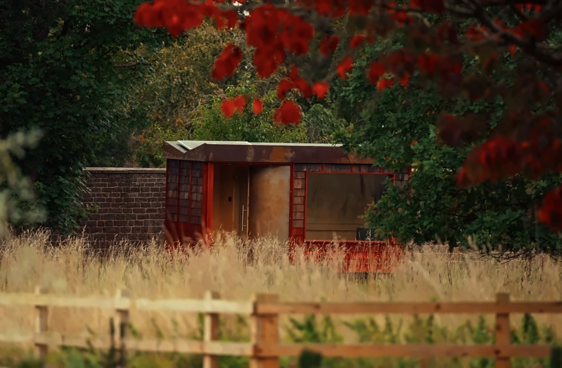A Rigg sauna sited in long grass beyond a timber fence, framed by autumn foliage